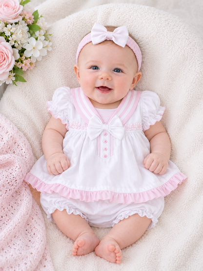 Baby in a pink and white outfit with a bow headband on a soft surface with flowers.