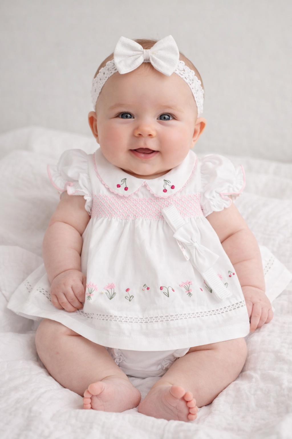 Baby wearing a white dress with pink embroidery and a matching headband on a white background
