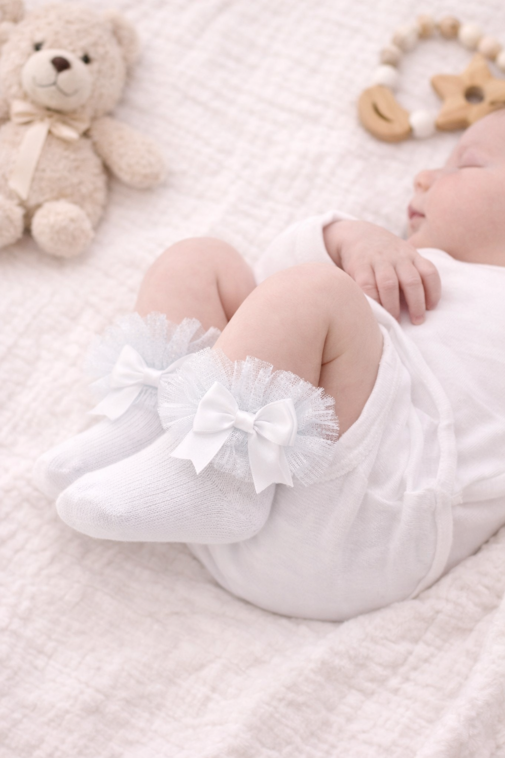 Flat lay mock-up of white frilly infant socks with satin bows, styled on a soft quilted blanket with neutral baby accessories including a teddy bear, wooden toys, and booties for a delicate nursery aesthetic.