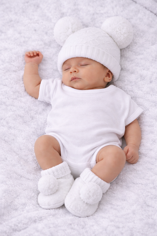 A sleeping newborn baby wearing a white short-sleeve bodysuit, a white knit hat with two pom-poms, and white booties, lying on a soft white blanket.