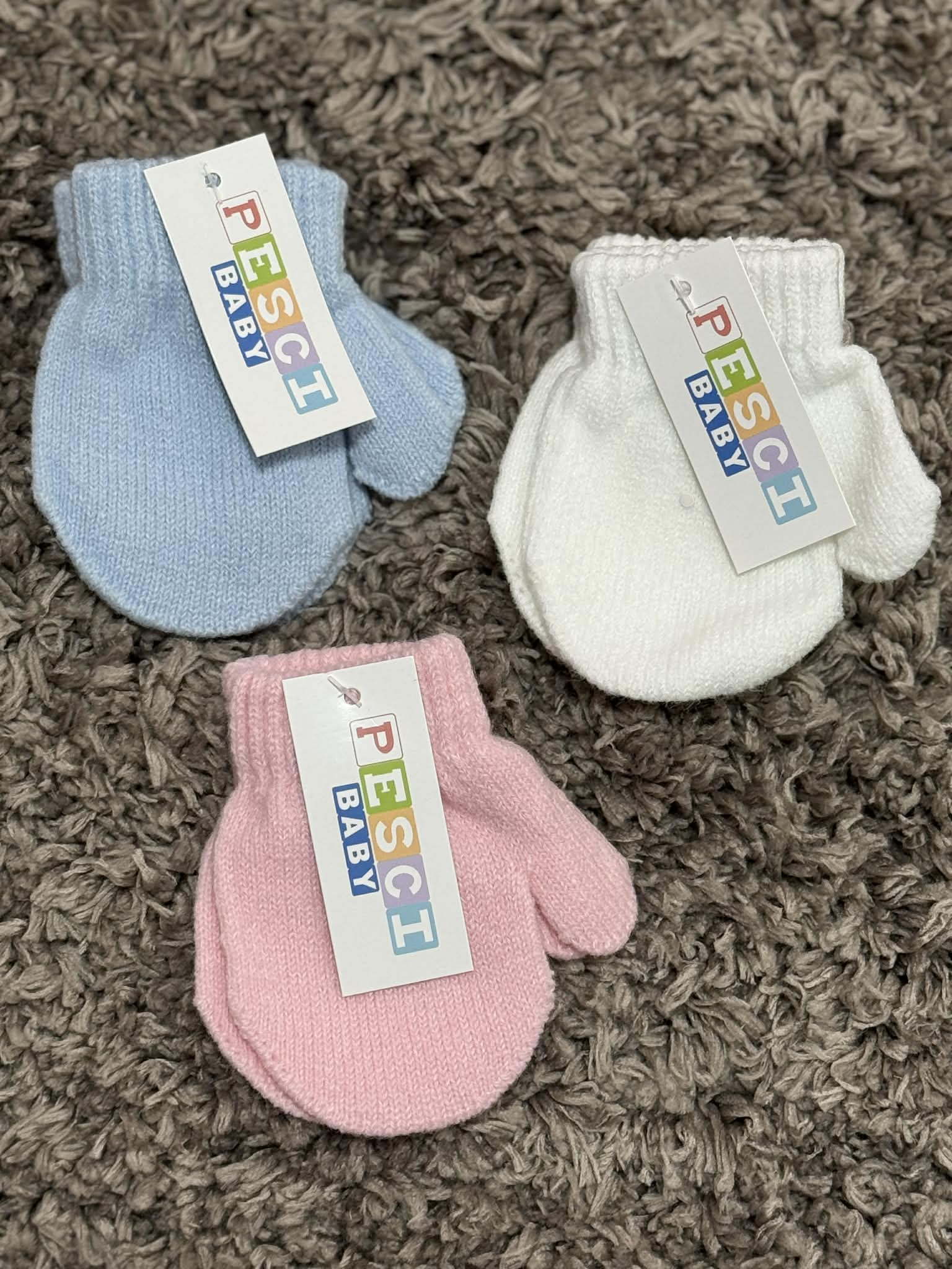 Three baby mittens in blue, pink, and white with 'PESCI BABY' labels on a textured brown surface.