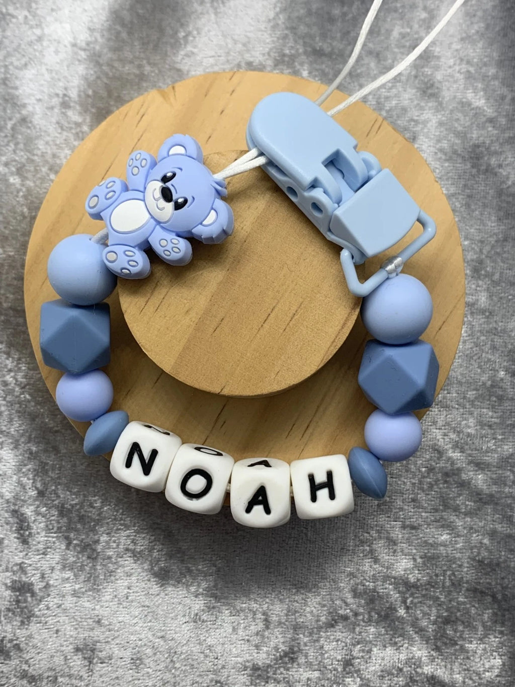 Baby teething clip with blue beads and a clip on a wooden board, featuring the name 'NOAH'. Personalisation and MAM Adapters Available.
