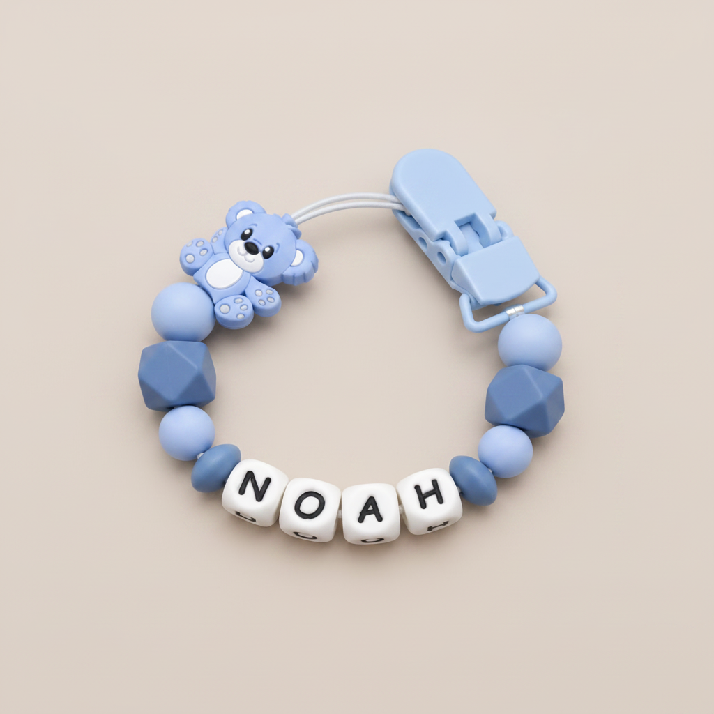 Blue baby dummy clip with a teddy bear design and 'NOAH' name on a beige background