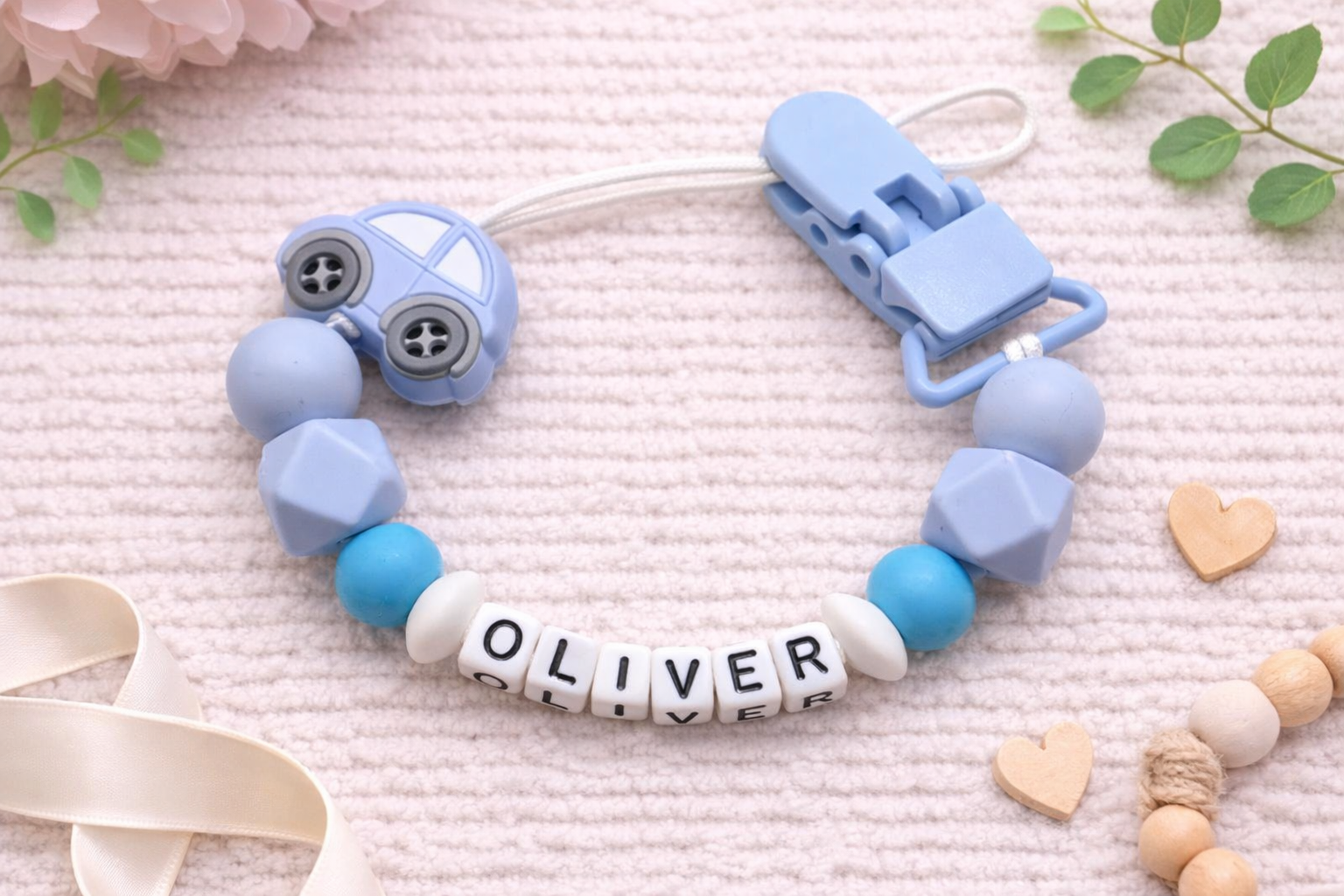 Baby dummy clip with blue and white beads and 'OLIVER' name tag on a soft pink background.