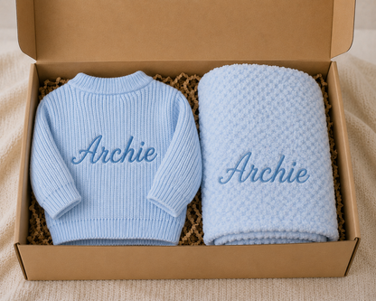 Blue baby sweater and blanket set with 'Archie' embroidery in a cardboard box.