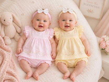 Two babies in pink and yellow outfits sitting on a soft surface with stuffed toys and flowers in the background.