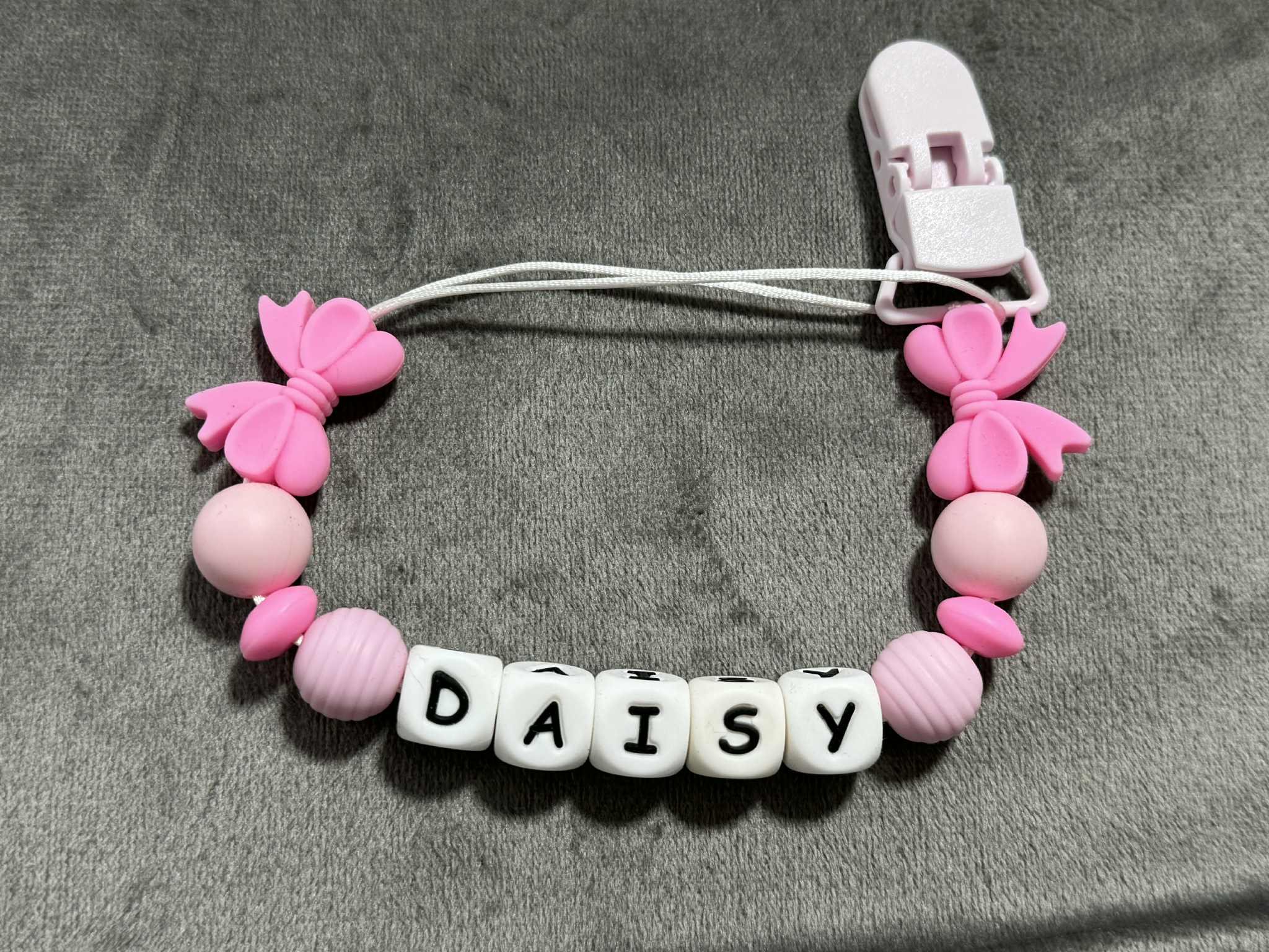 Pink pacifier clip with 'DAISY' letter beads on a gray background