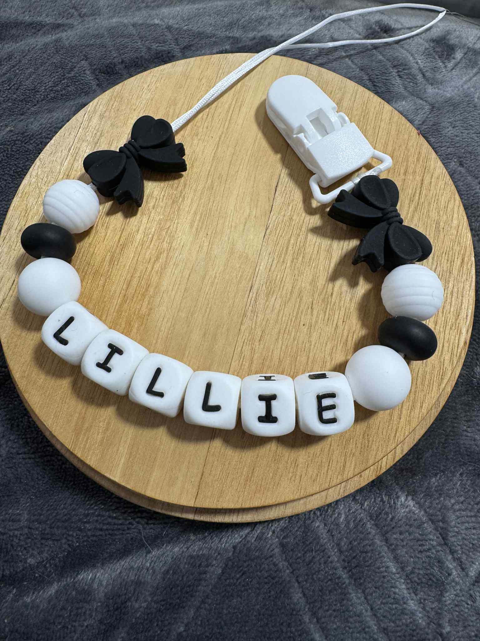 silicone dummy clip with black bows and white beads spelling 'LILLIE' on a wooden board.