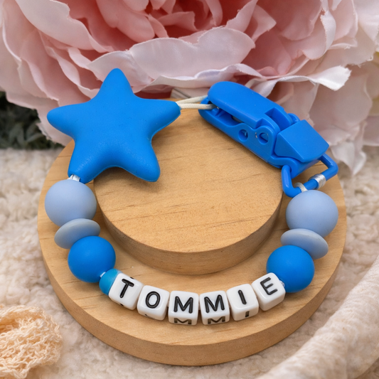 Blue and gray teething ring with 'TOMMIE' on a wooden base, pink flowers in the background