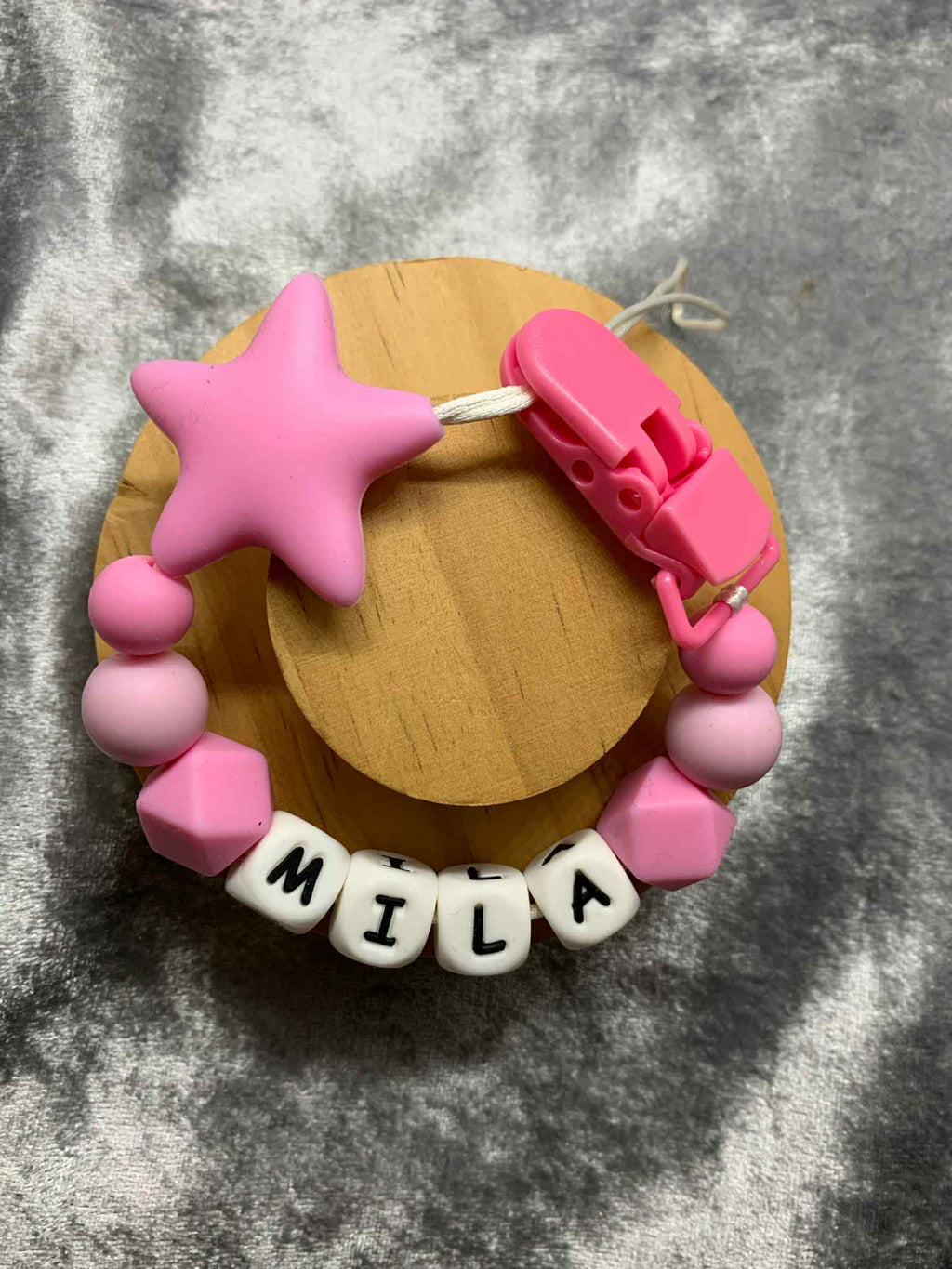 A beaded pacifier clip with bright pink beads, a pink star, and letter beads spelling "MILA" lying on a textured grey surface.