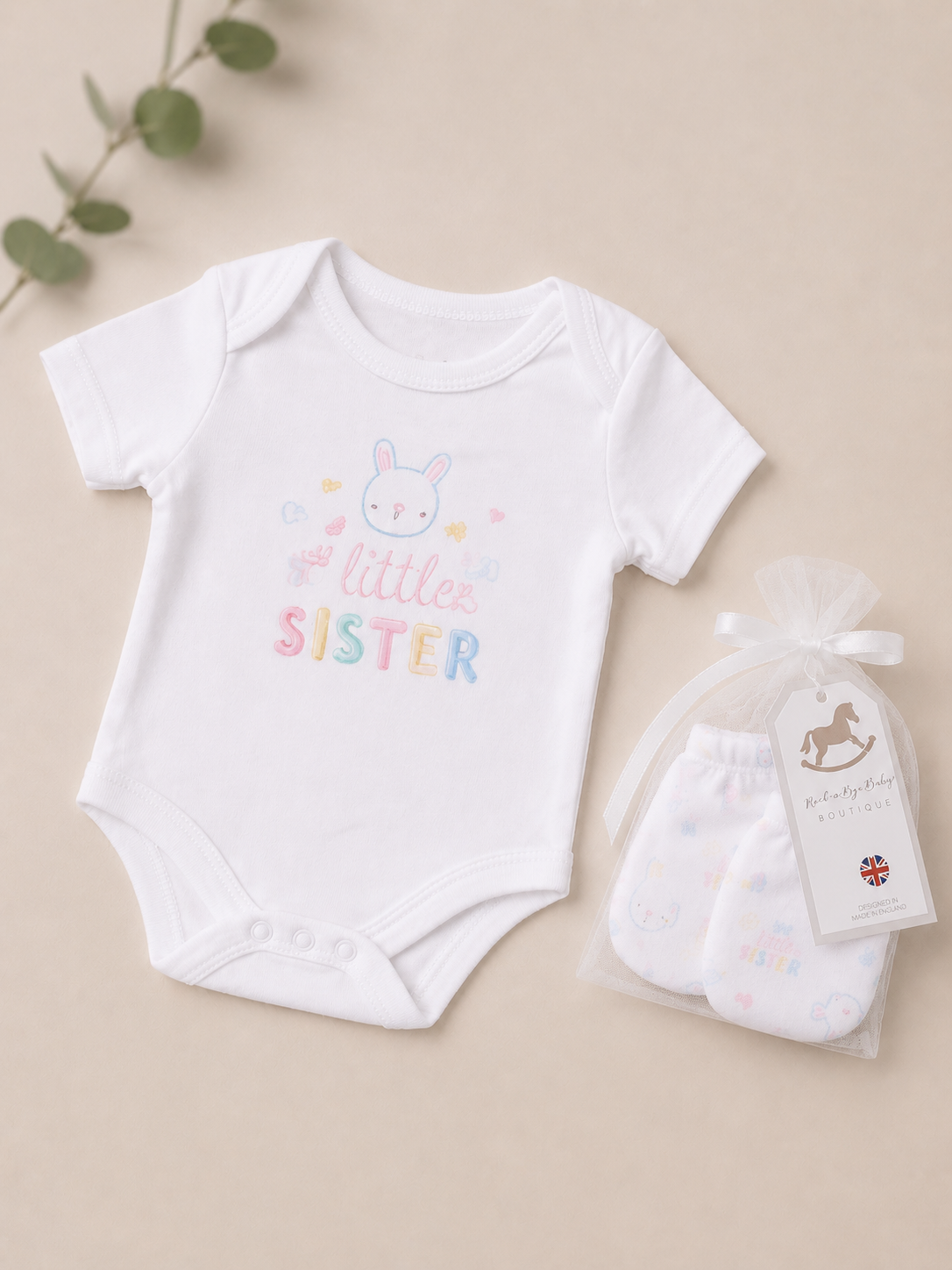 White baby onesie with 'Little Sister' text and a matching bag on a beige background