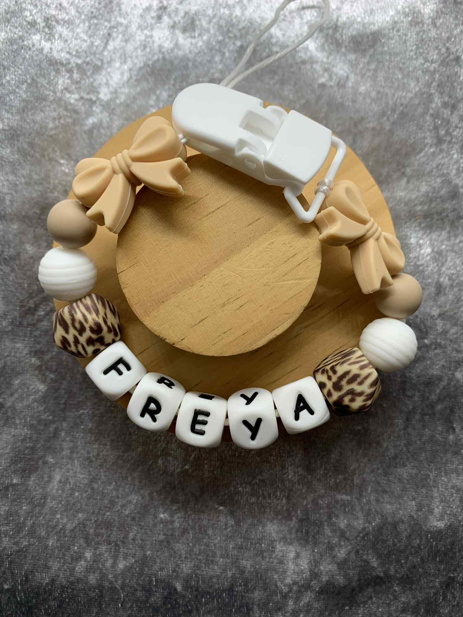 cream bow pacifier clip with decorative elements and 'FREYA' beads on a gray surface