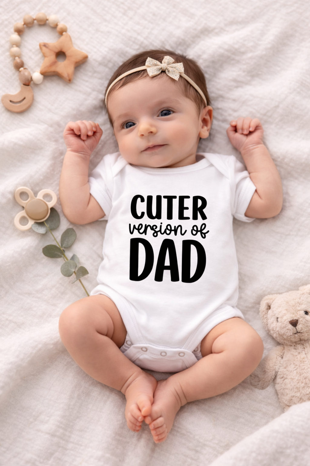 Baby wearing a onesie with 'Cuter version of Dad' text, surrounded by toys on a soft surface.