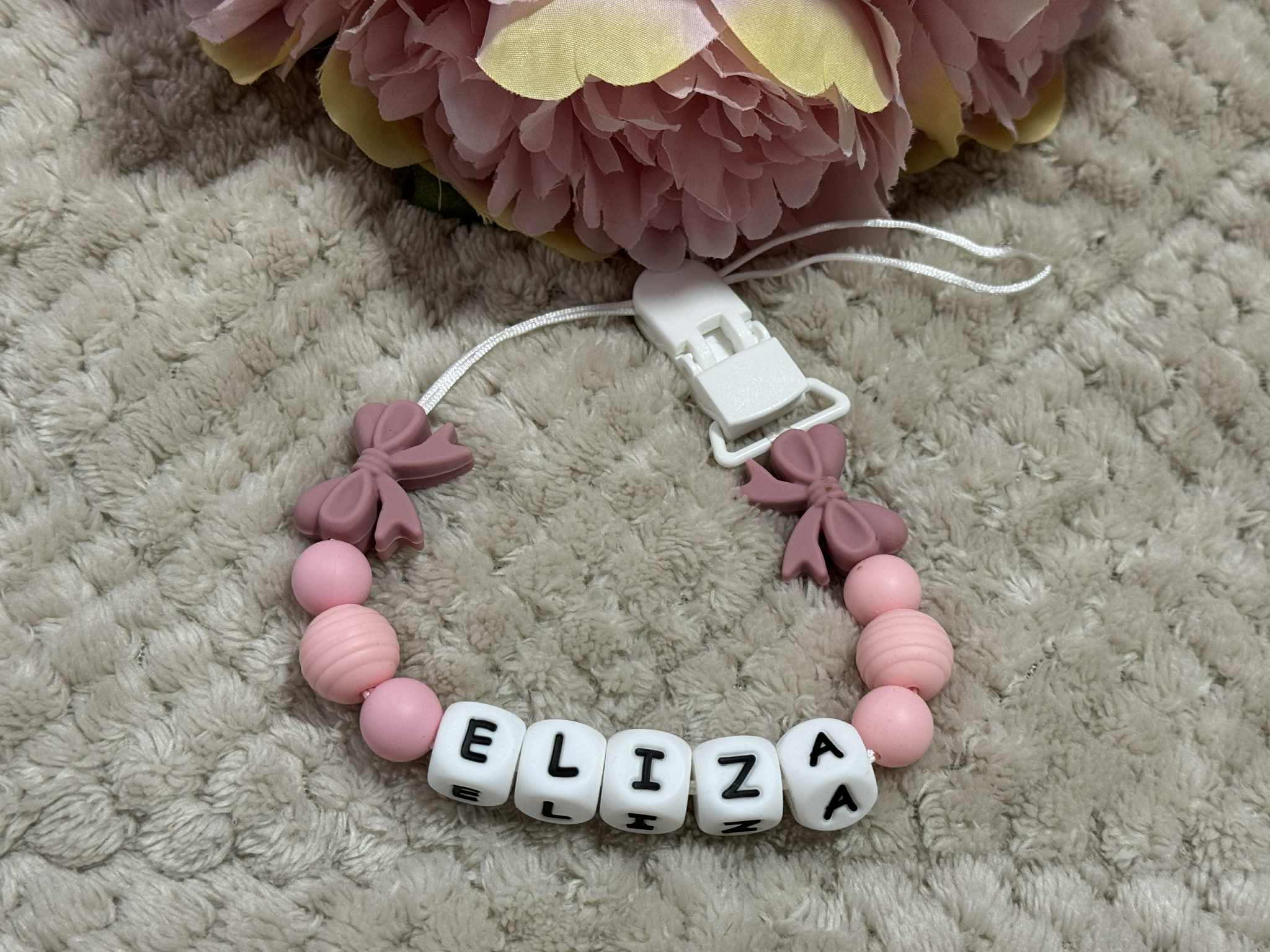 Pink baby teether dummy clip with 'ELIZA' beads on a textured surface with flowers in the background