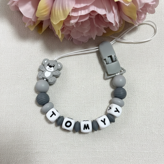 Personalised grey teddy bear dummy clip on a soft white luxury linen textured background.