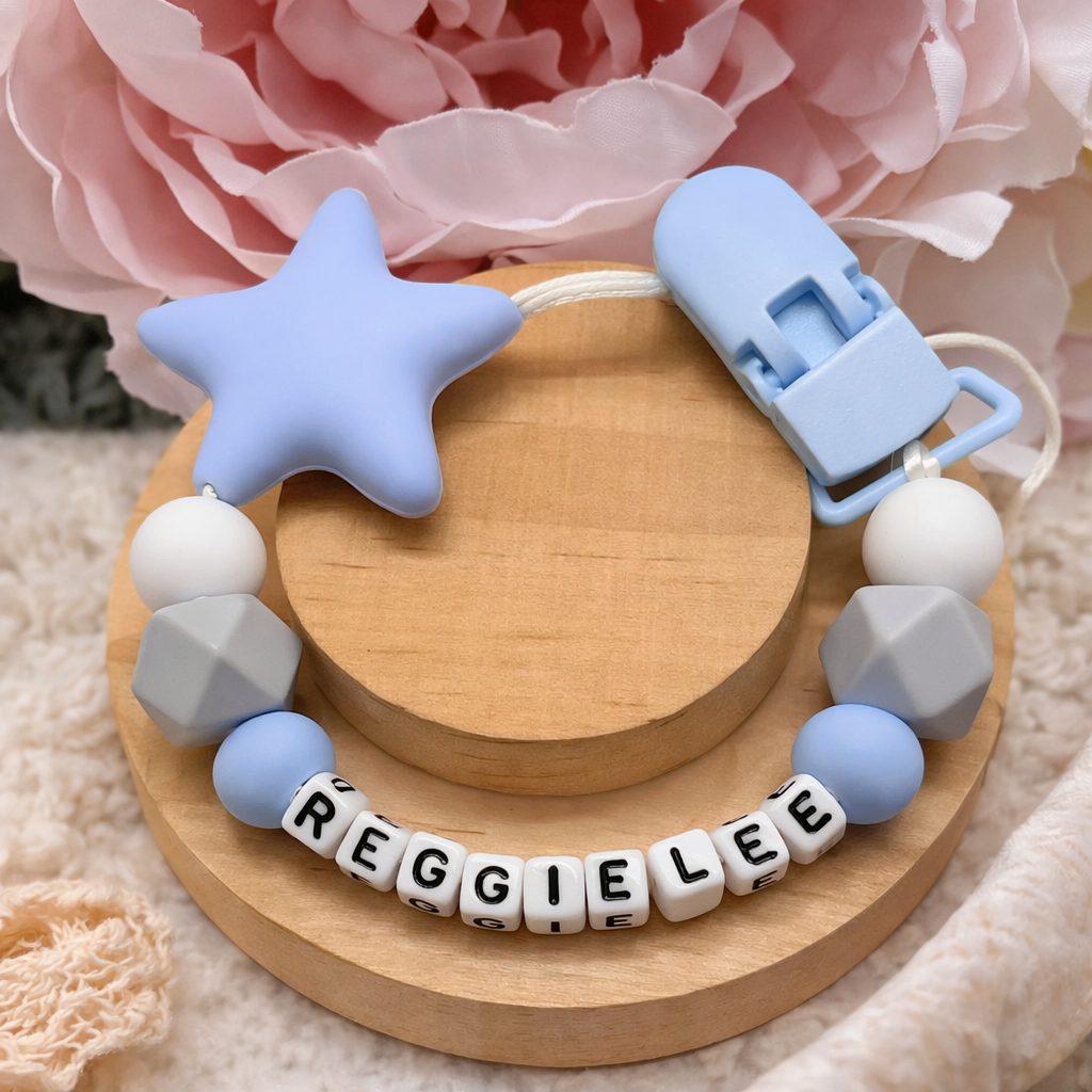 Baby teething necklace with star and geometric shapes on a wooden base, featuring the name 'REGGIE LEE'.