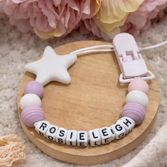 Baby dummy clip with name 'Rosie Leigh' on a wooden base with flowers in the background