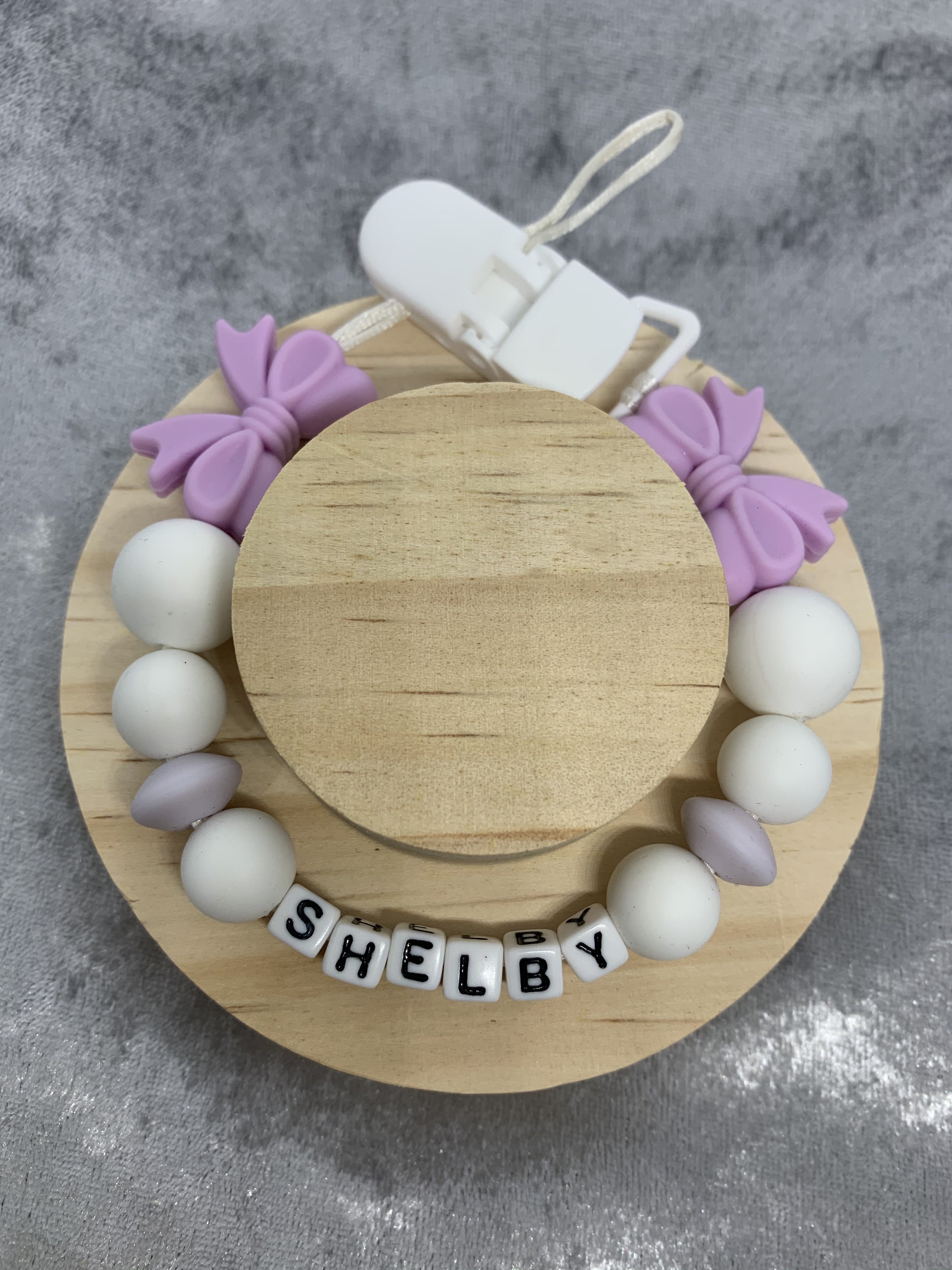 lilac bow dummy clip for babies with white beads, purple bows, and a clip on a gray background
