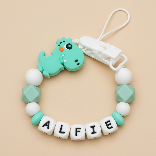 dummy clip with dinosaur design and 'ALFIE' beads on a beige background