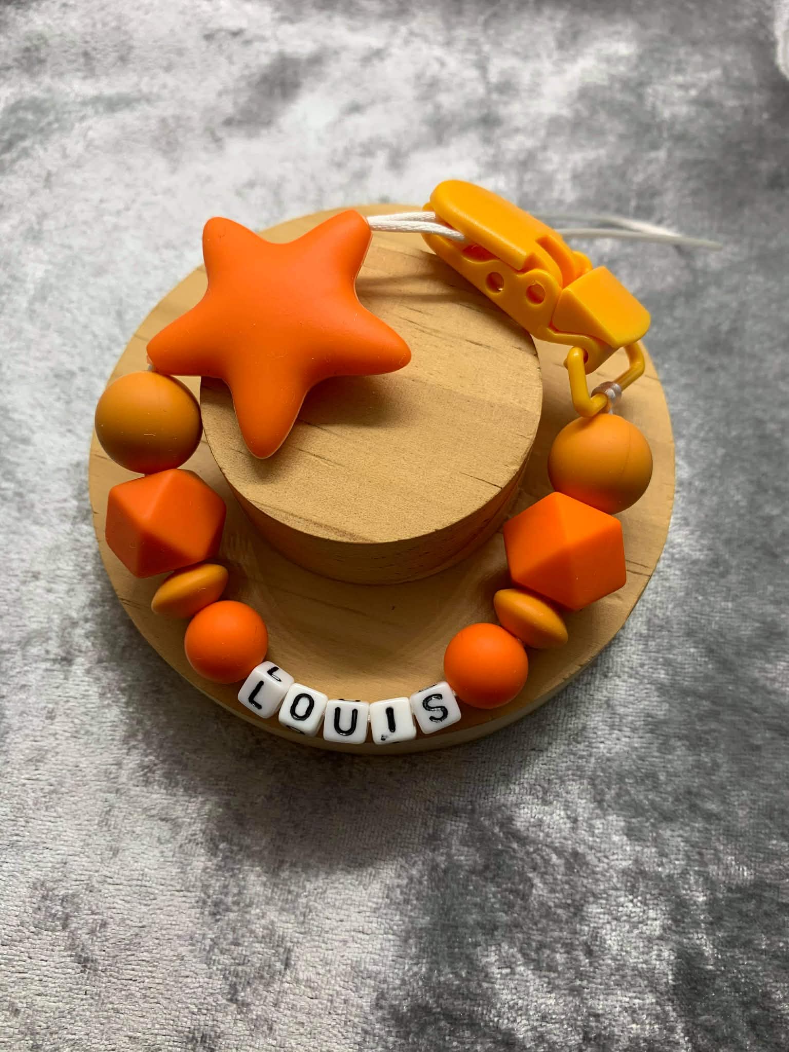 orange Silicone Dummy Clip with orange star personalised with the name 'Louis' on a grey textured surface. Can be personalised