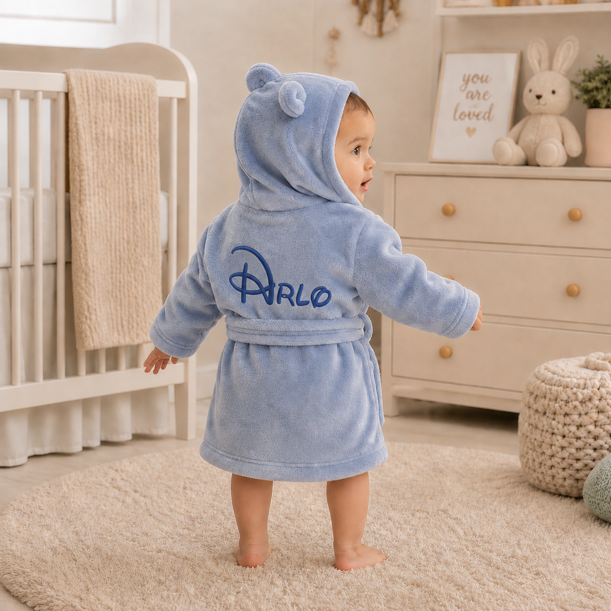 Baby in a blue robe with 'arlo' branding standing in a nursery.