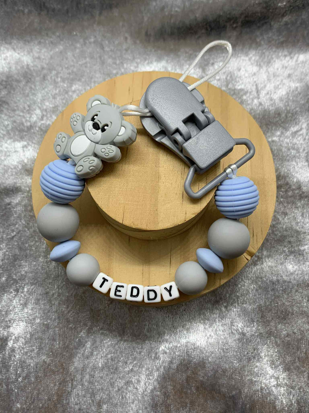Blue and grey teddybear dummy clip on a grey textured surface personalised with the name ''Teddy'. Personalisation and MAM Adapter options available.