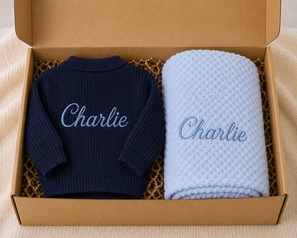 Navy sweater and light blue blanket with 'Charlie' embroidery in a cardboard box.
