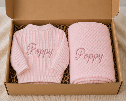 Pink baby sweater and blanket set with 'Poppy' embroidery in a cardboard box.