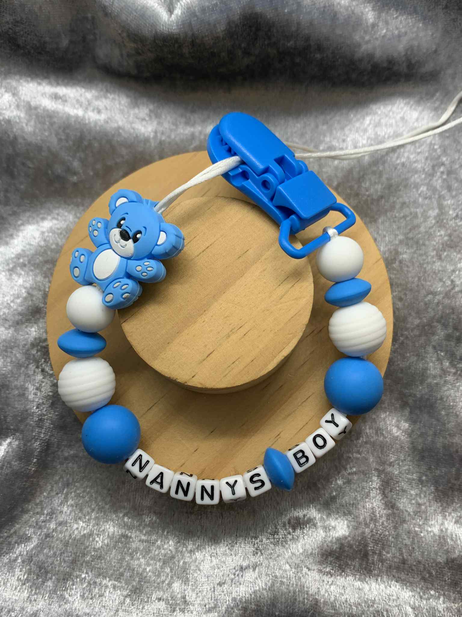 Blue teddybear dummy chain on a grey textured surface personalised with the name 'Nanny's Boy'. Personalisation and MAM Adapter options available.