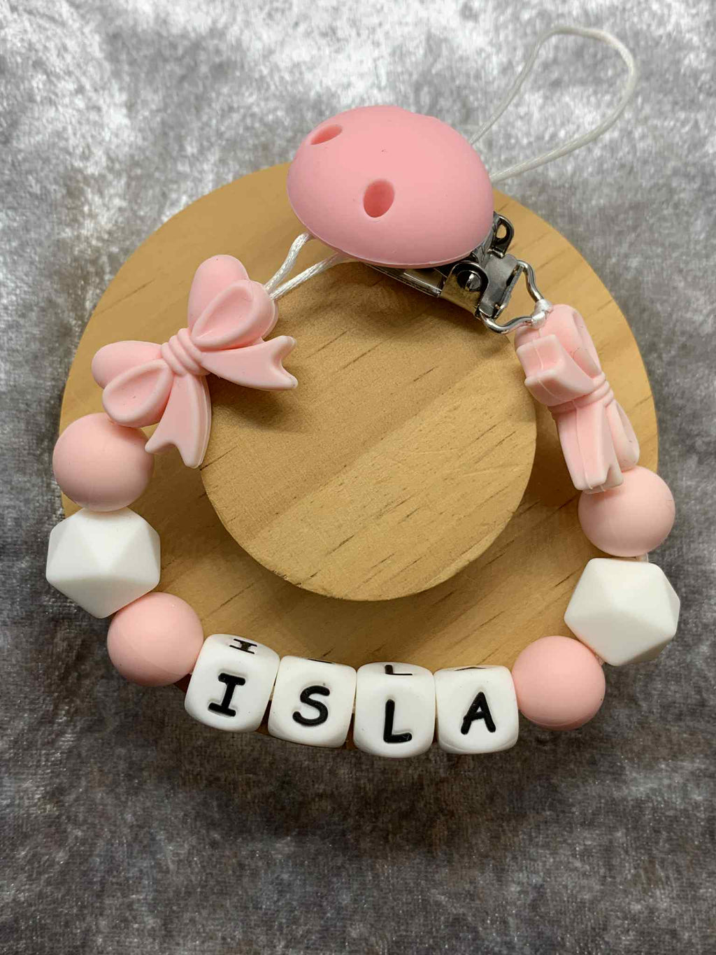 Pacifier clip with pink and white beads spelling 'ISLA' on a wooden base.