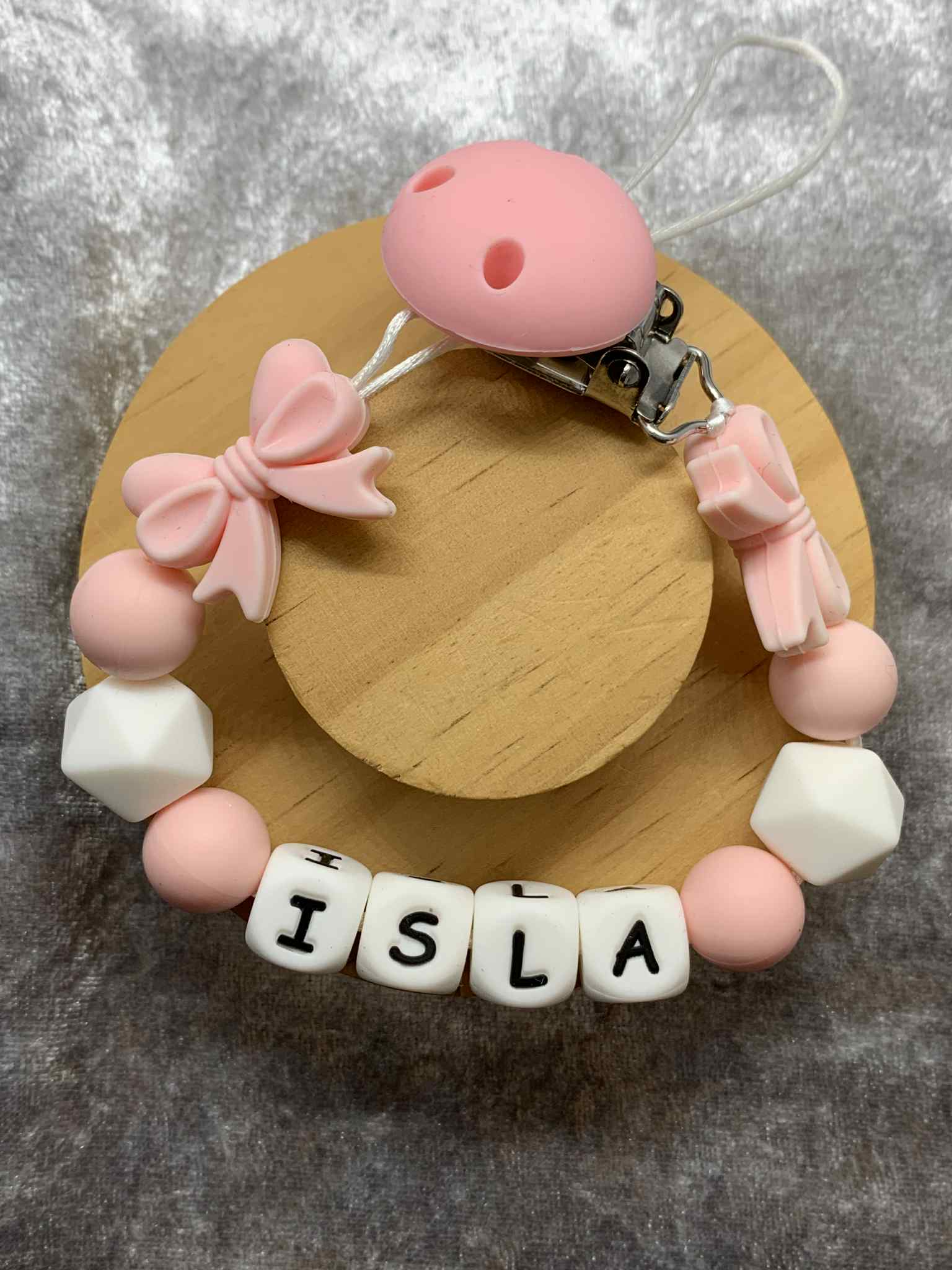 Pacifier clip with pink and white beads spelling 'ISLA' on a wooden base.