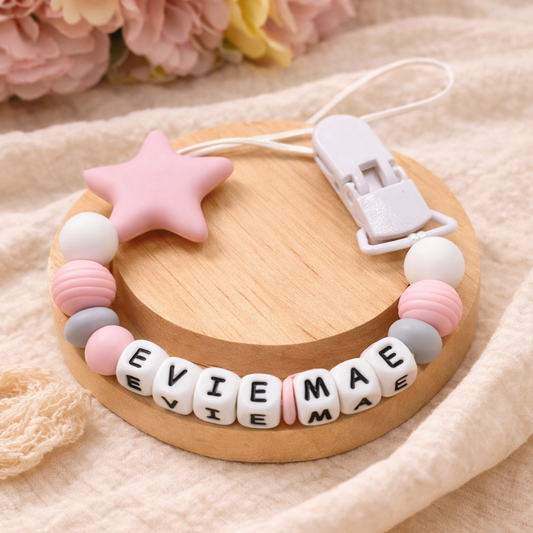 Baby teething necklace with name beads on a wooden base, surrounded by flowers.