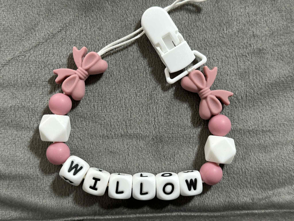 pink pacifier holder with pink bow charms and white beads spelling 'WILLOW' on a gray fabric background.