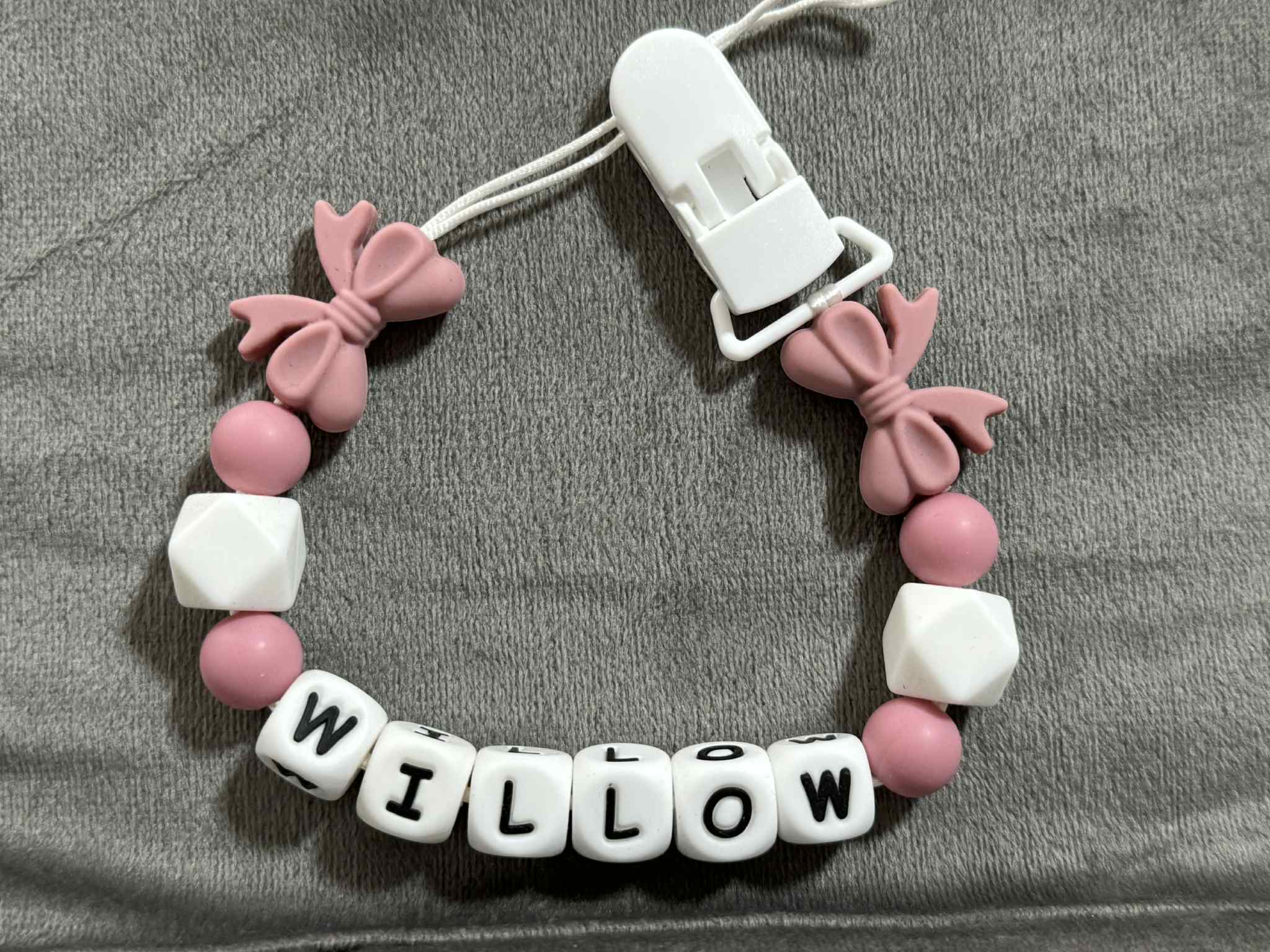 pink pacifier holder with pink bow charms and white beads spelling 'WILLOW' on a gray fabric background.