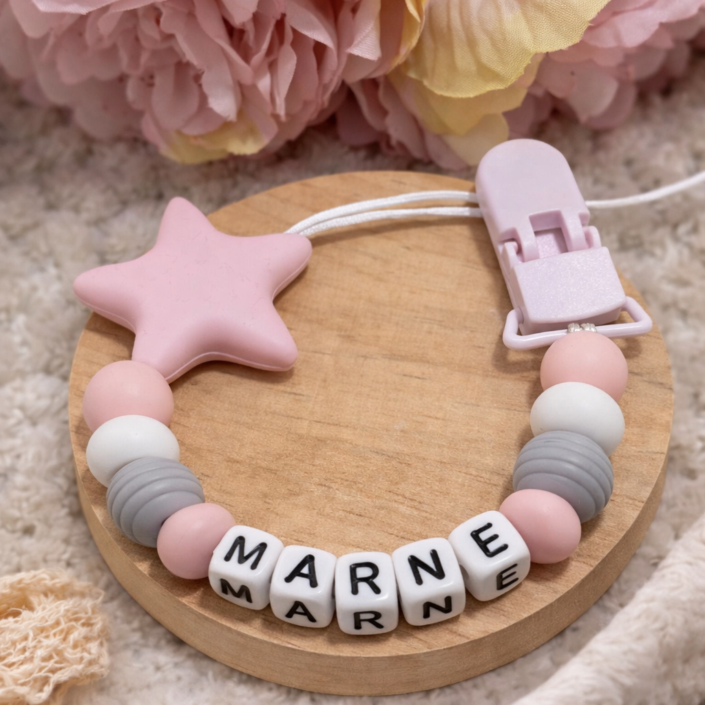 Baby pacifier clip with 'MARNIE' on beads, pink star, and clip on wooden base with flowers in background