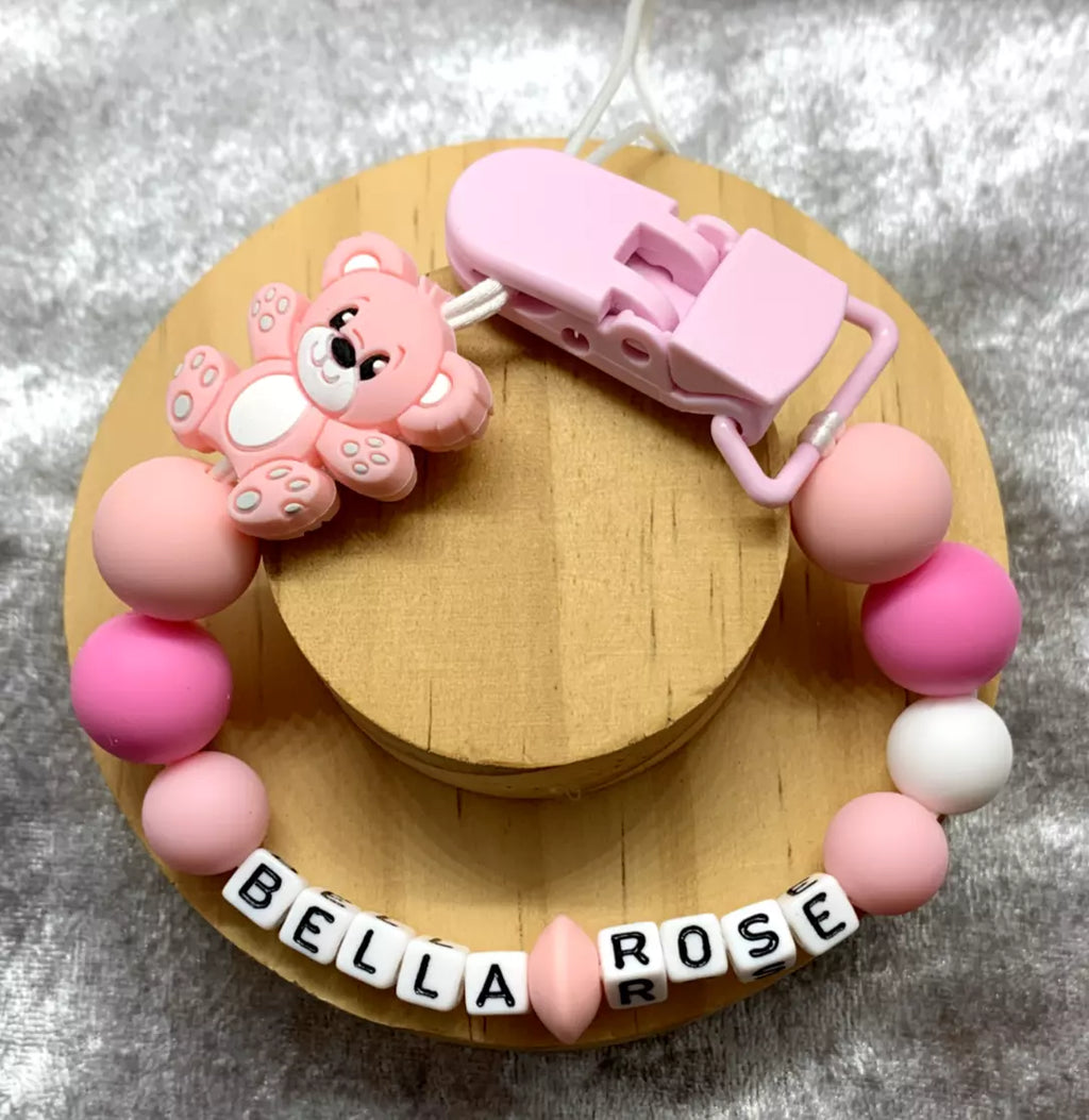 Pink teddybear dummy clip on a grey textured surface. Personalised with the name 'Bella-Rose'. Change of name is available in the personalisation box. MAM Adapters are available too.