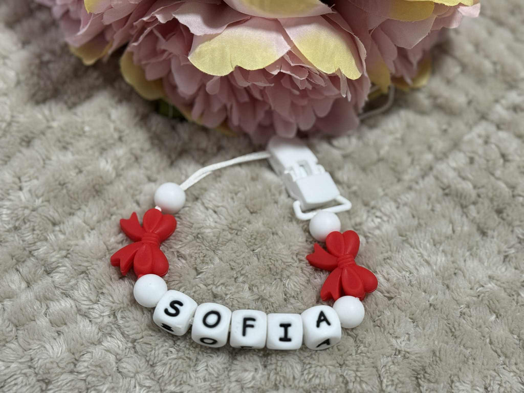pacifier clip with 'SOFIA' beads and red flower charms on a textured surface with flowers in the background