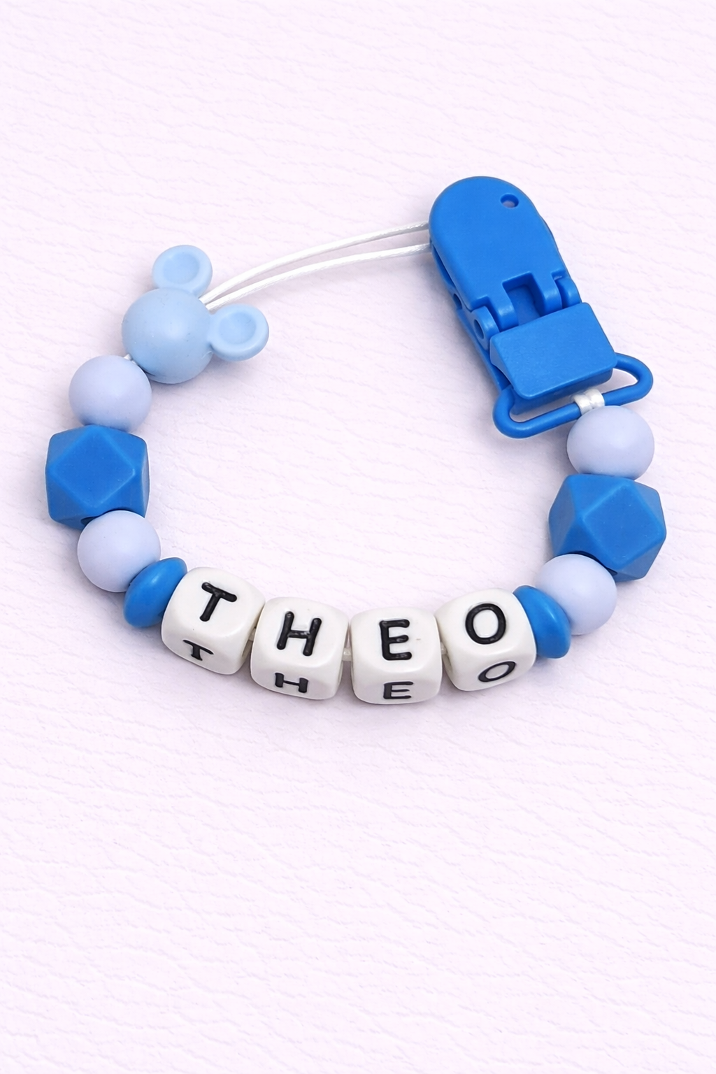 Blue and white pacifier clip with letter beads spelling 'THEO' on a light purple background