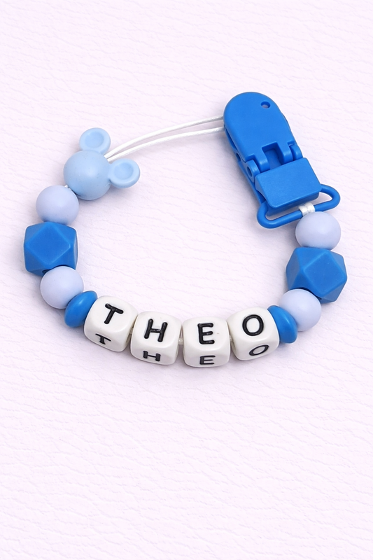 Blue and white pacifier clip with letter beads spelling 'THEO' on a light purple background