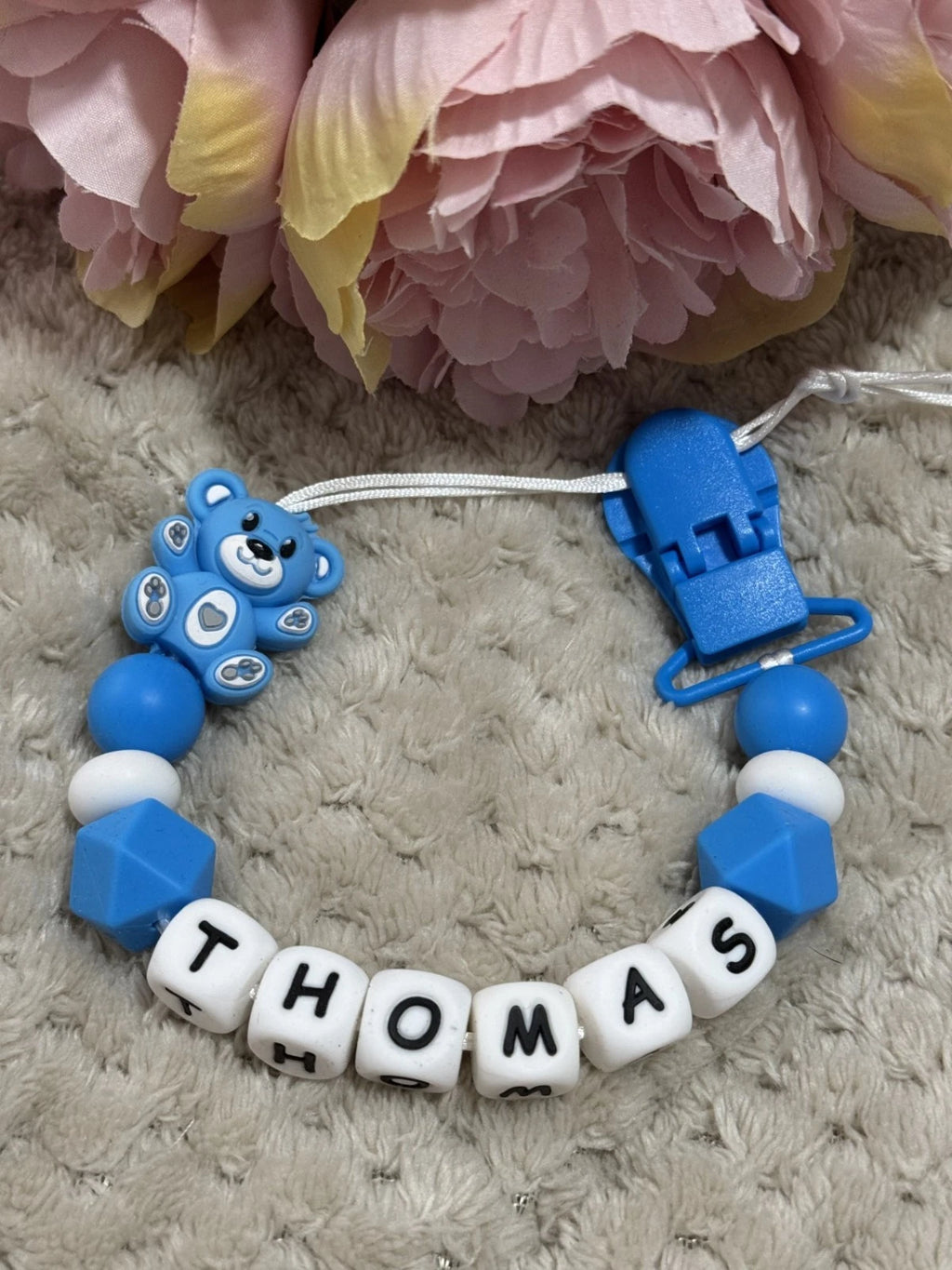 Blue and white baby soother clip with 'THOMAS' on a textured surface with pink flowers in the background