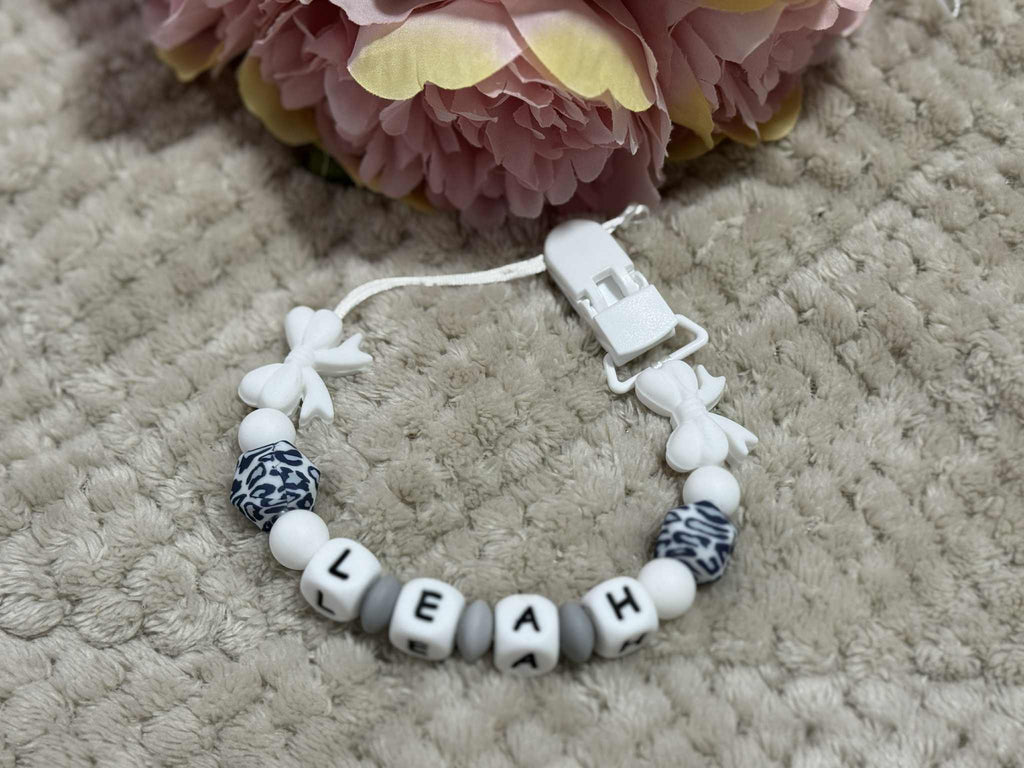 Beaded pacifier clip with 'LEAH' on a textured surface with pink flowers in the background